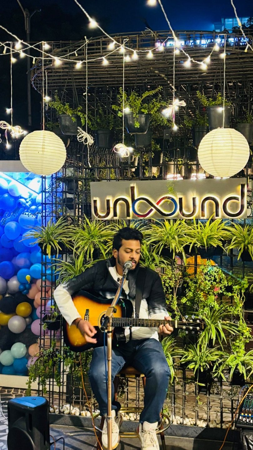 A singer with a guitar in front of the lit UnBound sign and the green wall, paper lanterns above